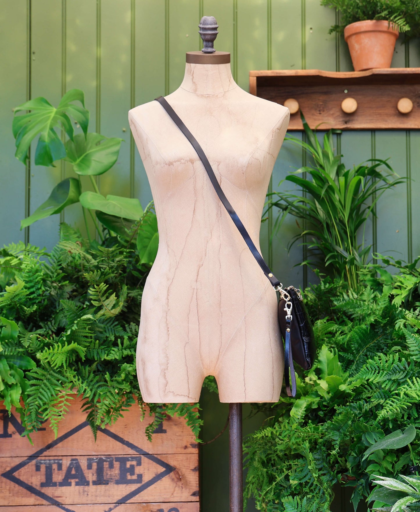 A beige mannequin torso wearing a black croc print crossbody bag with a long strap, set against a green foliage and wooden wall backdrop.