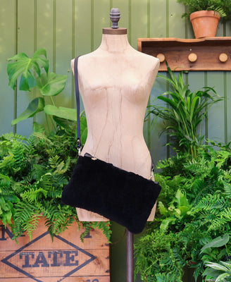 A black sheepskin and leather cross body bag with a thin black strap, displayed on a beige dress form mannequin. The mannequin is set against a background of green potted plants and a green wooden panelled wall.